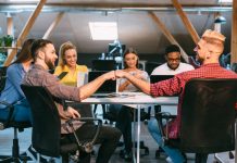 Two team members trusting each other while shaking hands while seated at a conference table surrounded by colleagues in a contemporary office environment.